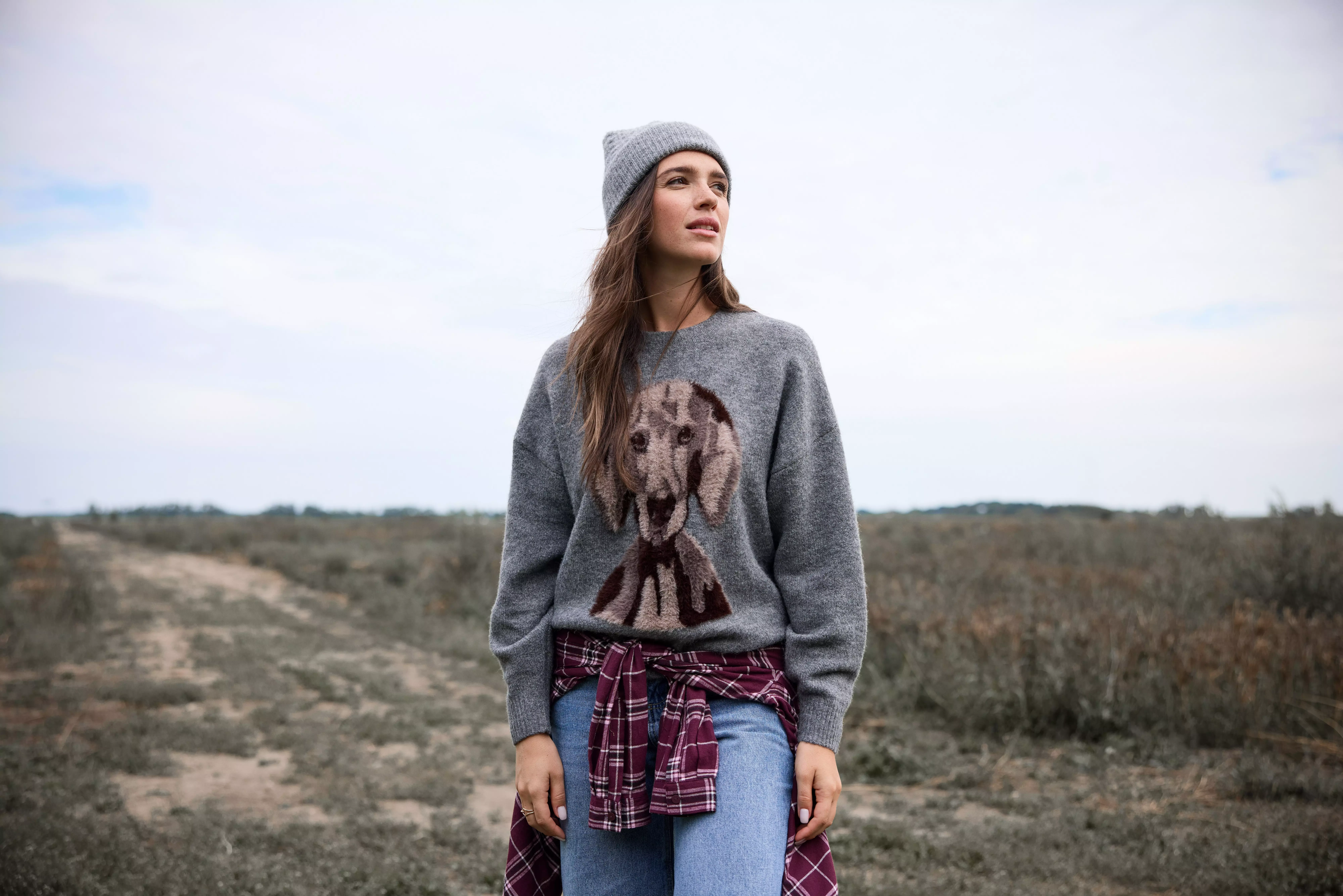 Pepco woman wearing grey knit jumper with dog motif, beanie hat and jeans outdoors.